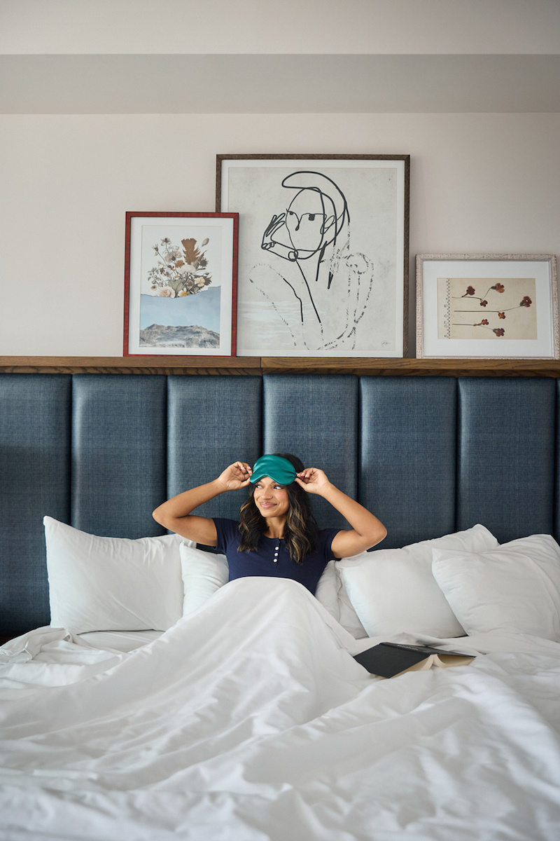 A woman sitting up in a bed with a navy headboard, wearing a sleep mask and holding a book.