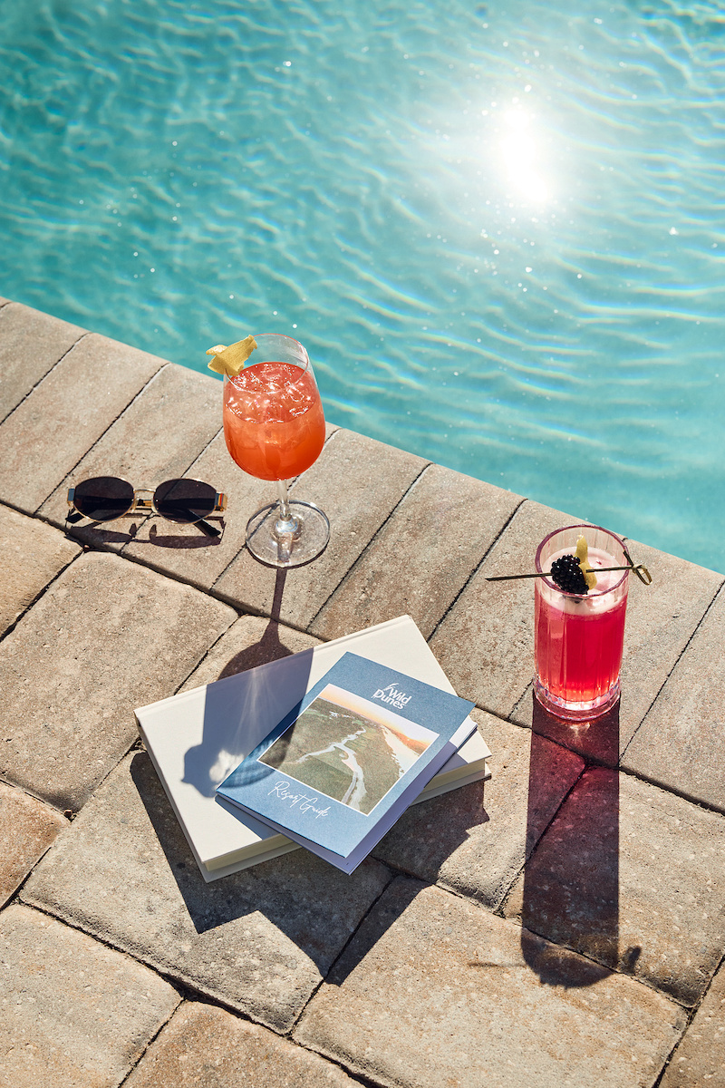 Two glasses of red cocktails, a book, and sunglasses on a stone pool deck next to a swimming pool.