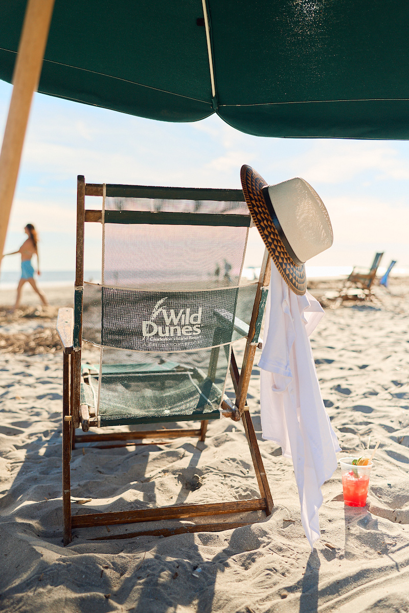 A beach chair with the Wild Dunes logo, a straw hat, and a white towel under a green umbrella on the sand.