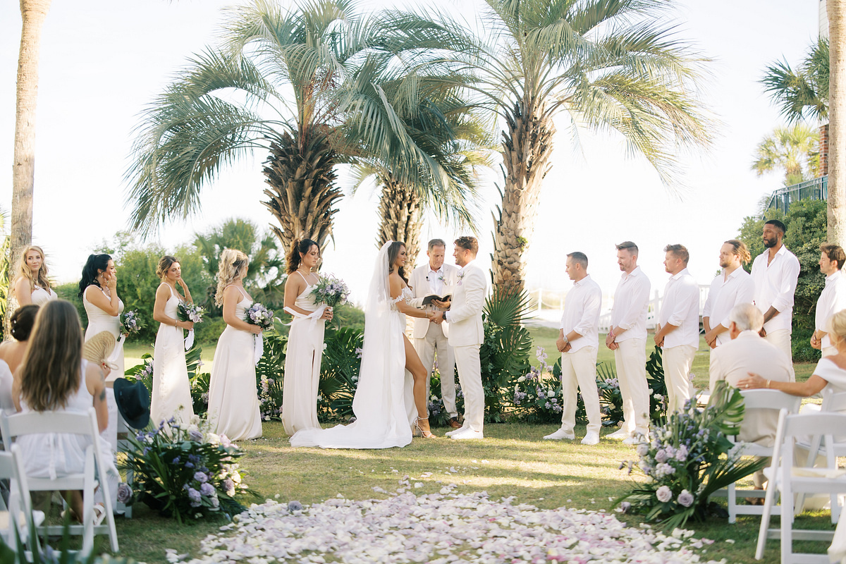 A bride and groom exchange vows at an outdoor wedding ceremony, flanked by a bridal party and groomsmen under palm trees.
