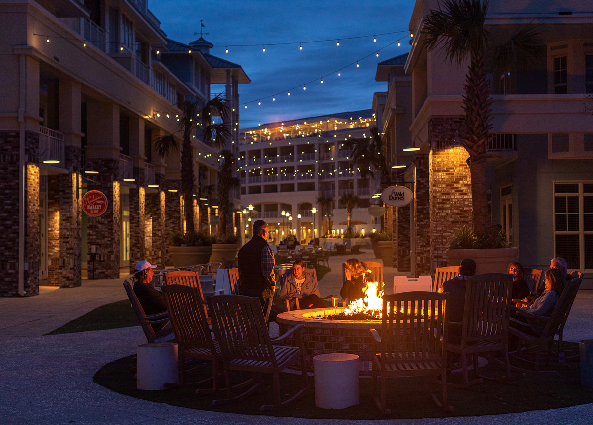 People sitting in rocking chairs around a lit outdoor fire pit at dusk, surrounded by resort buildings with string lights.