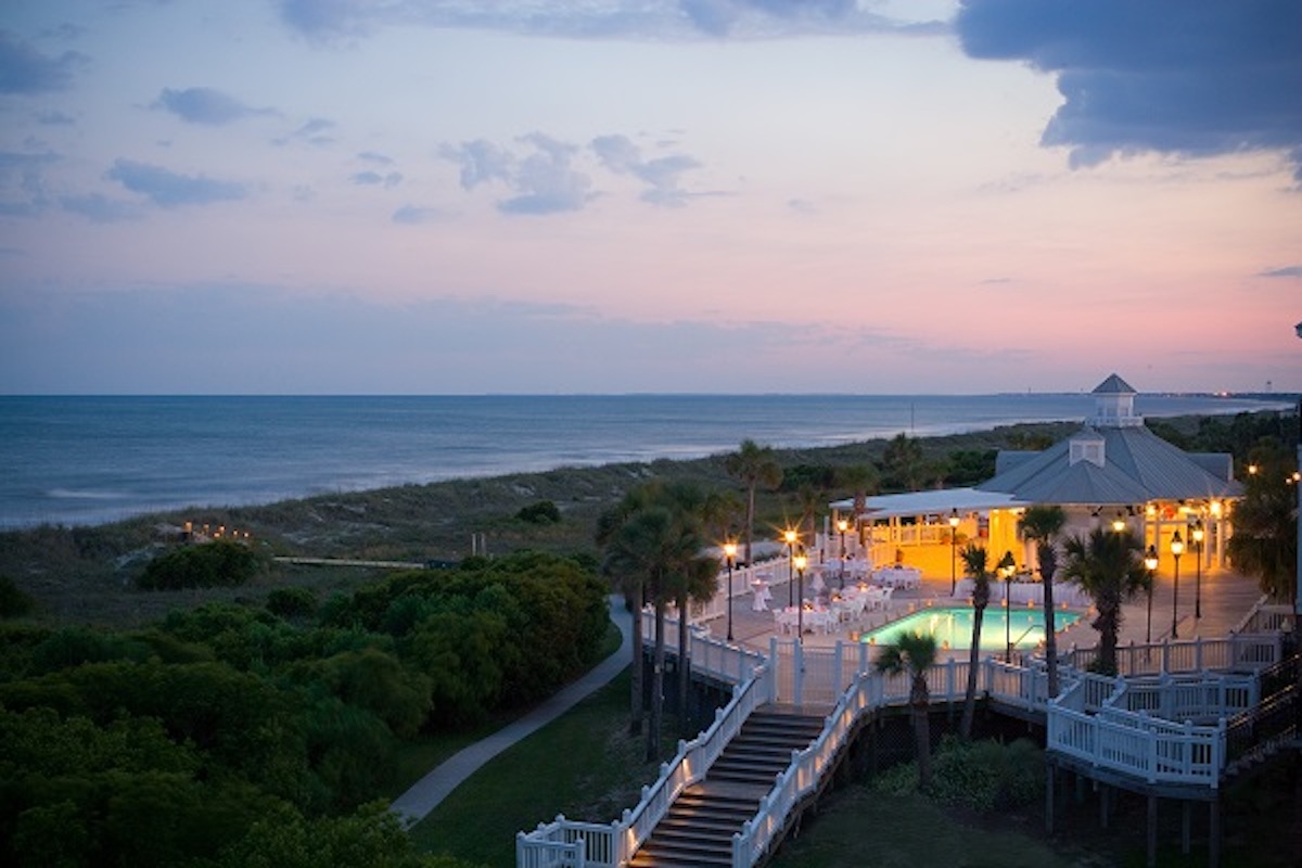 An oceanfront resort at twilight, featuring an illuminated pool and building beside a beach.