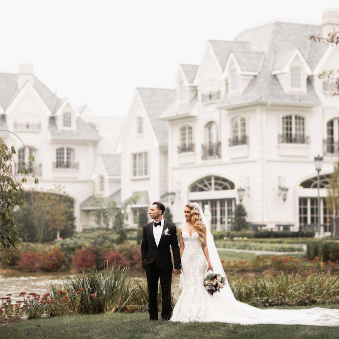 A bride and groom stand together in front of a large, elegant mansion with gardens and a pond. The groom wears a black suit, and the bride is in a white gown holding a bouquet.