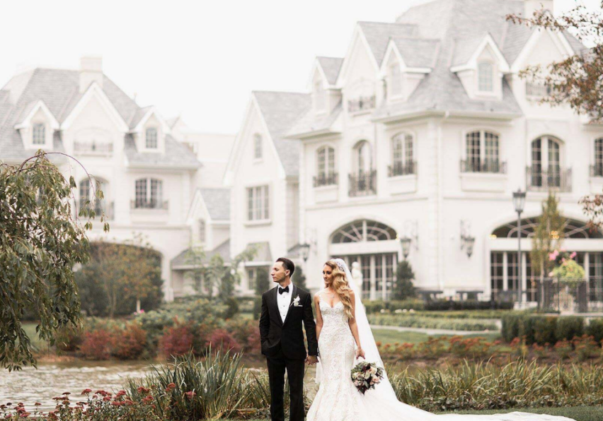 A bride and groom stand together in front of a large, elegant mansion with gardens and a pond. The groom wears a black suit, and the bride is in a white gown holding a bouquet.