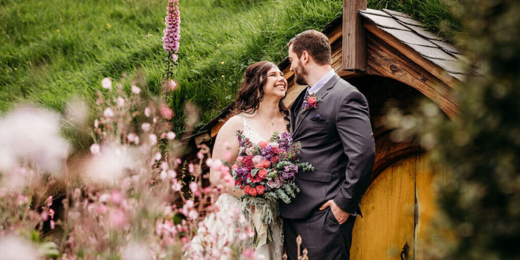 A bride and groom stand closely together, smiling, in front of a small wooden door surrounded by greenery and flowers. The bride holds a large bouquet.
