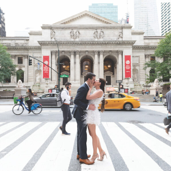 A couple kisses on a crosswalk in front of an ornate building, with pedestrians, cyclists, and a yellow taxi nearby.