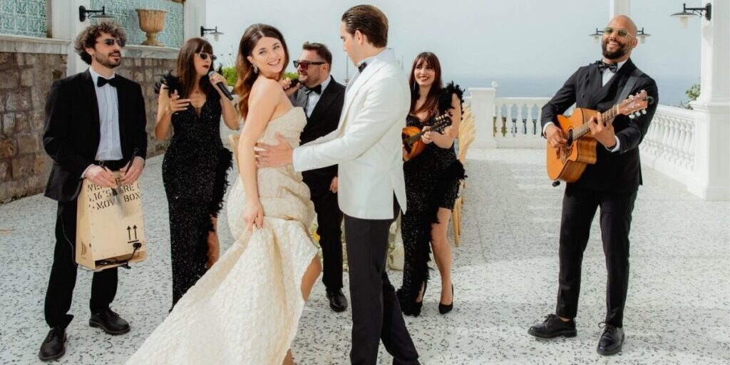 A bride and groom dance outdoors while a band with guitars and singers performs nearby; the scene appears festive and lively.