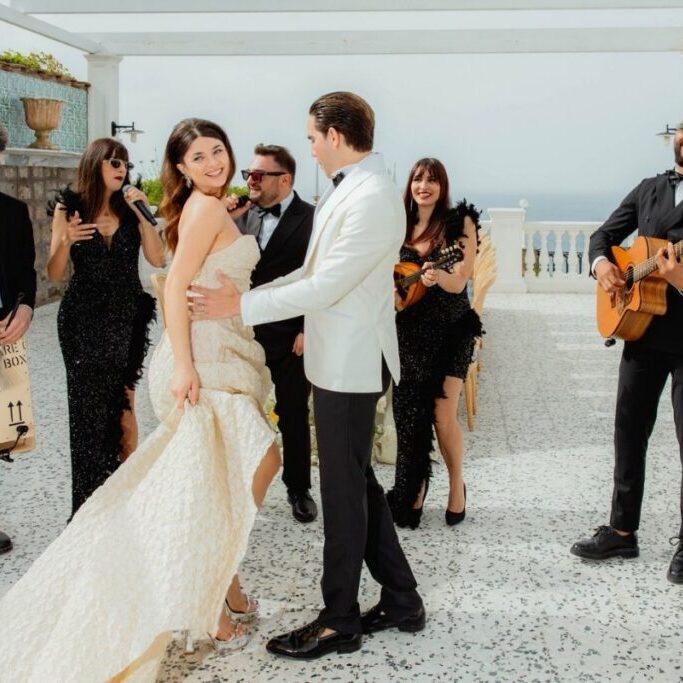 A bride and groom dance outdoors while a band with guitars and singers performs nearby; the scene appears festive and lively.