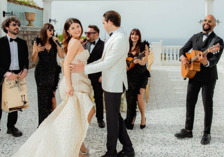 A bride and groom dance outdoors while a band with guitars and singers performs nearby; the scene appears festive and lively.