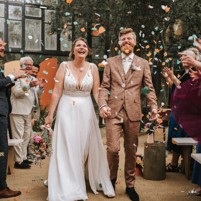 A bride and groom walk down the aisle smiling as guests throw confetti in celebration at an indoor wedding ceremony.
