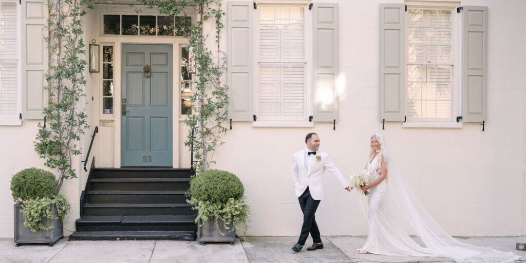 A bride and groom stand hand in hand on a sidewalk in front of a light-colored building with green shutters and a green door.