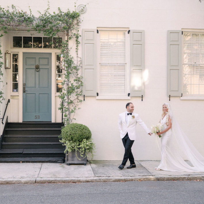 A bride and groom stand hand in hand on a sidewalk in front of a light-colored building with green shutters and a green door.