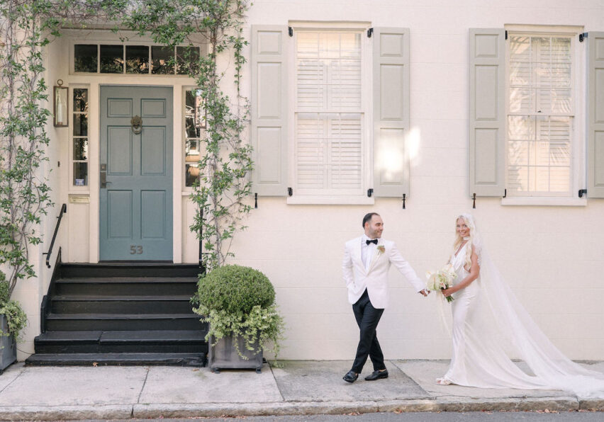 A bride and groom stand hand in hand on a sidewalk in front of a light-colored building with green shutters and a green door.