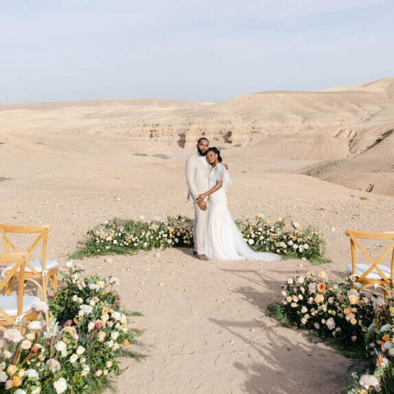 a couple embraces at the altar surrounded by a semi circle of white and pink florals with desert mountains in the background