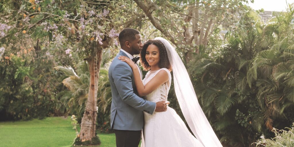 Bride and groom pose together outdoors; the groom wears a blue suit, and the bride wears a white dress with a long veil. Lush greenery and trees appear in the background.