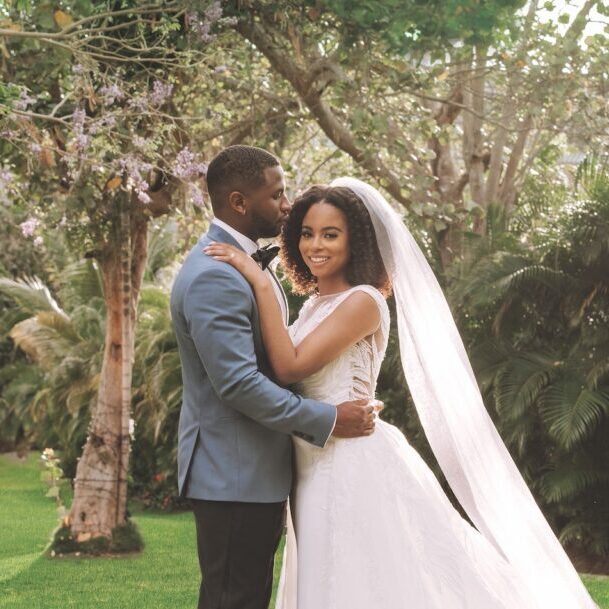 Bride and groom pose together outdoors; the groom wears a blue suit, and the bride wears a white dress with a long veil. Lush greenery and trees appear in the background.