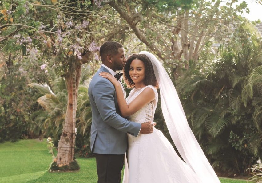 Bride and groom pose together outdoors; the groom wears a blue suit, and the bride wears a white dress with a long veil. Lush greenery and trees appear in the background.
