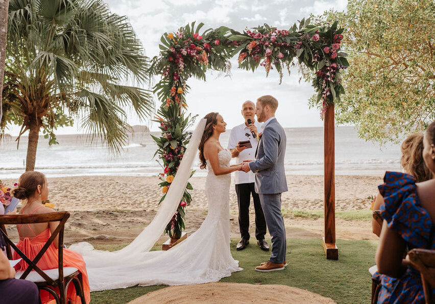 A bride and groom exchange vows under a floral arch at a beachside wedding ceremony, with guests seated nearby and the ocean in the background.