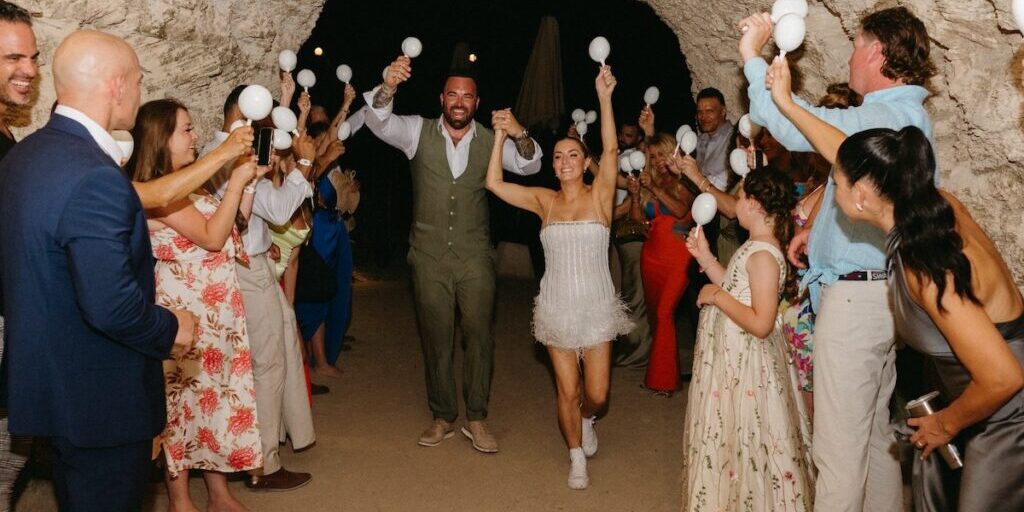 A couple walks hand in hand through a tunnel as guests celebrate, holding white maracas and clapping.