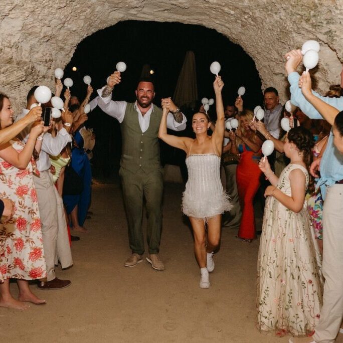 A couple walks hand in hand through a tunnel as guests celebrate, holding white maracas and clapping.
