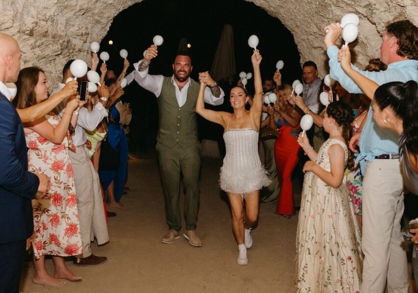 A couple walks hand in hand through a tunnel as guests celebrate, holding white maracas and clapping.