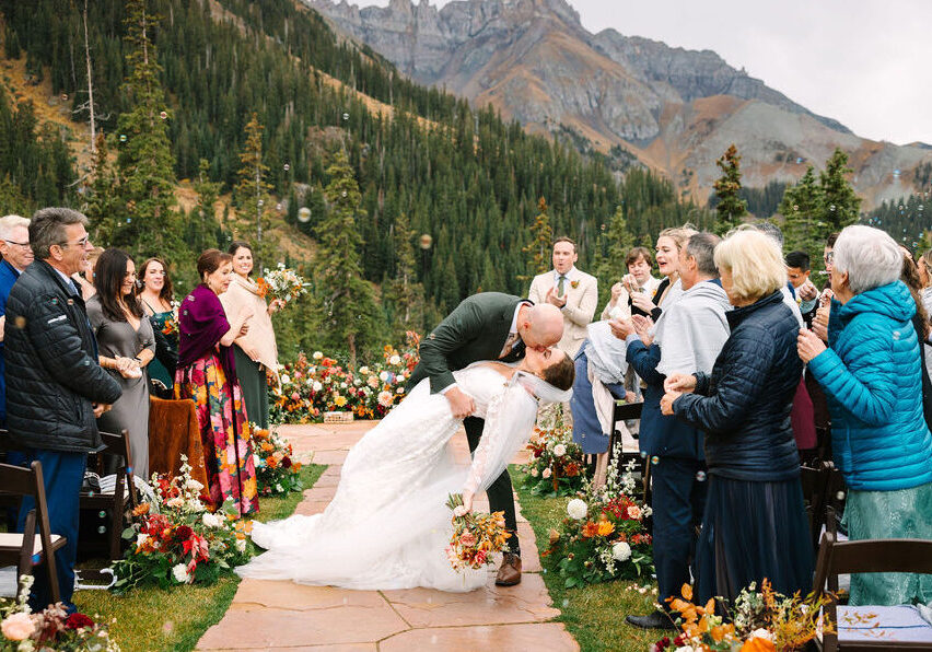 A bride and groom kiss at an outdoor mountain wedding ceremony, surrounded by guests and autumn floral arrangements on a stone path.