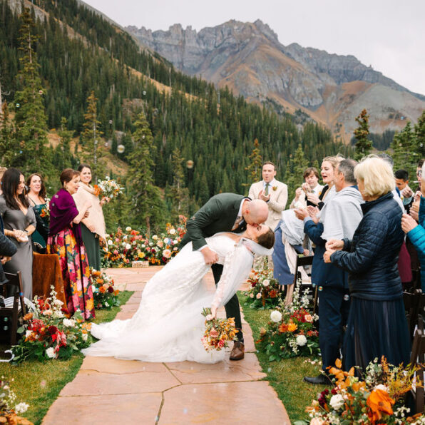 A bride and groom kiss at an outdoor mountain wedding ceremony, surrounded by guests and autumn floral arrangements on a stone path.