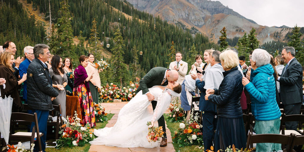 A bride and groom kiss at an outdoor mountain wedding ceremony, surrounded by guests and autumn floral arrangements on a stone path.