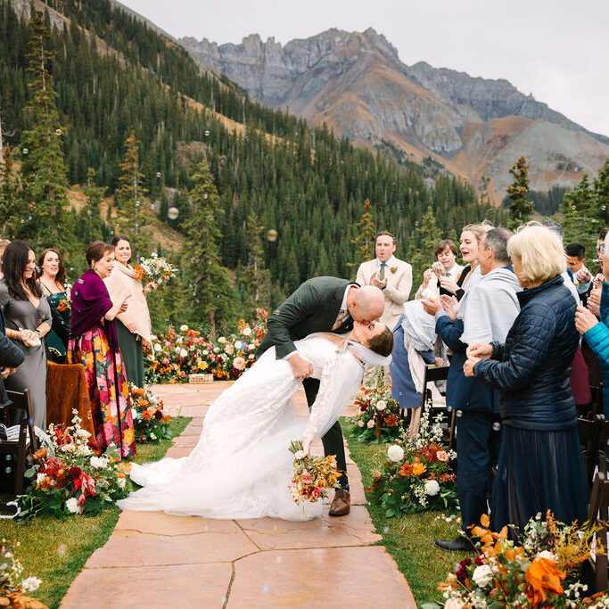 A bride and groom kiss at an outdoor mountain wedding ceremony, surrounded by guests and autumn floral arrangements on a stone path.