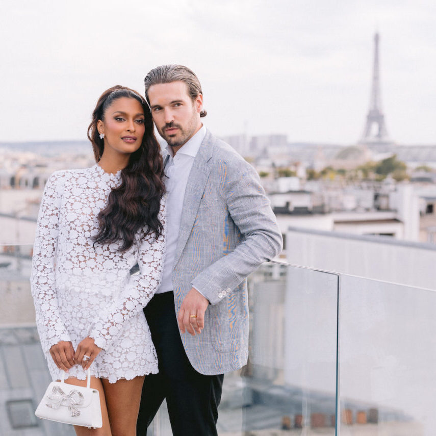 A woman in a white dress and a man in a grey blazer pose together on a rooftop with the Eiffel Tower visible in the background.