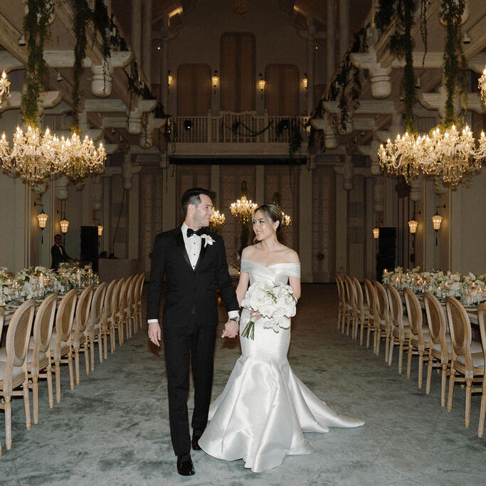 A bride and groom walk hand in hand down an aisle in an elegantly decorated hall with chandeliers and long banquet tables.