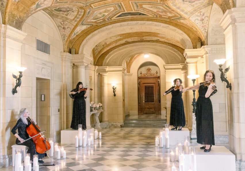 Four women in black dresses perform with string instruments in an ornate hall with arched ceilings, columns, and candles on the checkered floor.