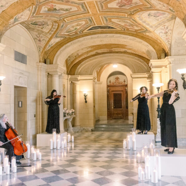 Four women in black dresses perform with string instruments in an ornate hall with arched ceilings, columns, and candles on the checkered floor.