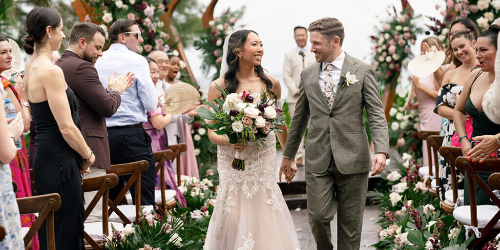 A bride and groom walk down the aisle together, smiling, surrounded by guests clapping and floral decorations at an outdoor wedding ceremony.
