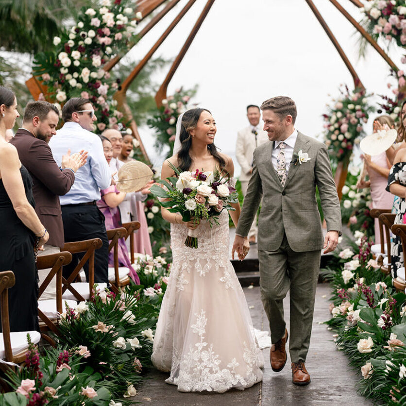 A bride and groom walk down the aisle together, smiling, surrounded by guests clapping and floral decorations at an outdoor wedding ceremony.