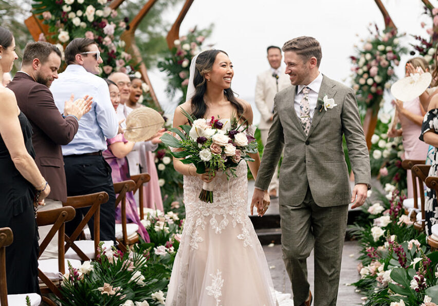A bride and groom walk down the aisle together, smiling, surrounded by guests clapping and floral decorations at an outdoor wedding ceremony.
