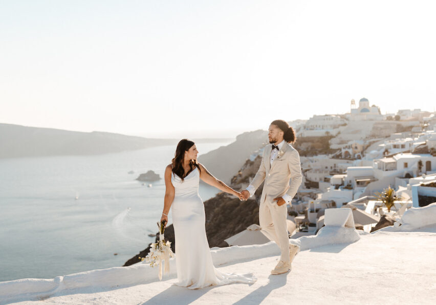 A bride and groom hold hands and walk on a white terrace overlooking the sea and white buildings, likely in Santorini, Greece, under bright sunlight.