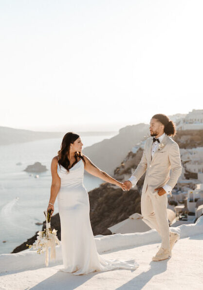 A bride and groom hold hands and walk on a white terrace overlooking the sea and white buildings, likely in Santorini, Greece, under bright sunlight.