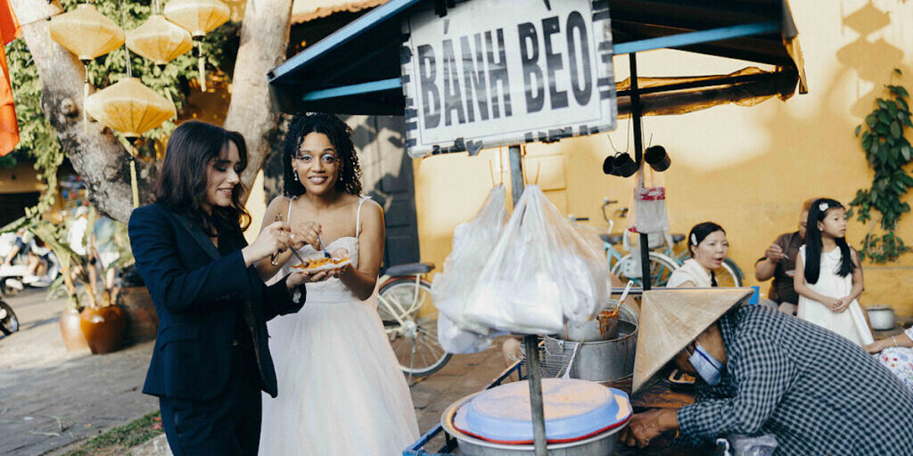 Two women, one in a suit and one in a white dress, eat food at a Bánh Bèo street food stall while a vendor in a conical hat serves them.