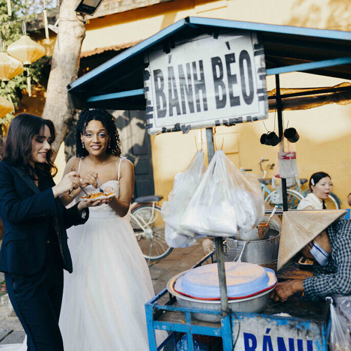 Two women, one in a suit and one in a white dress, eat food at a Bánh Bèo street food stall while a vendor in a conical hat serves them.