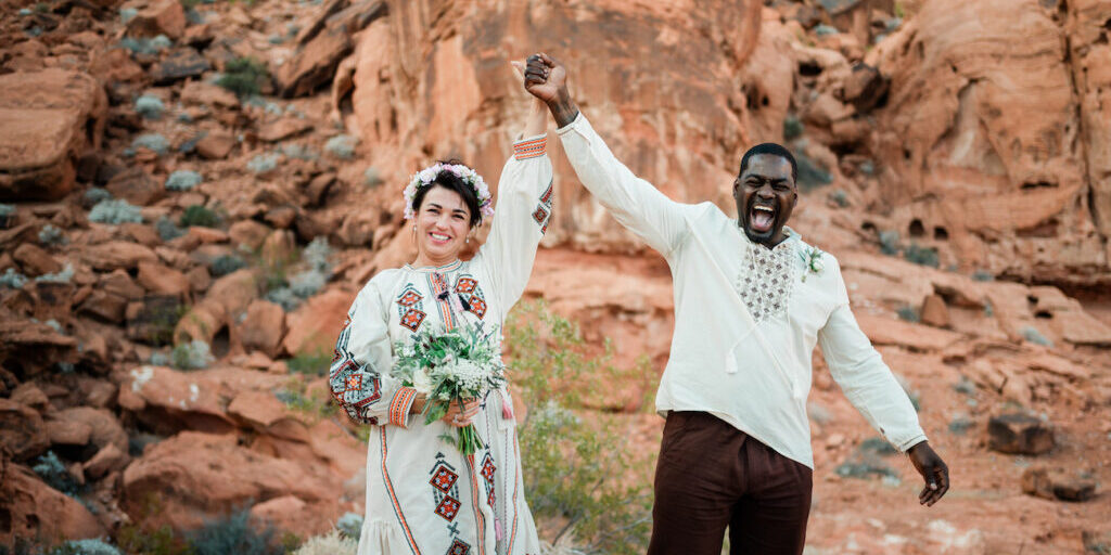 A smiling couple in embroidered clothing stands in a rocky desert landscape, holding hands up in celebration. The woman holds a bouquet and wears a flower crown.