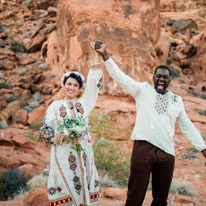 A smiling couple in embroidered clothing stands in a rocky desert landscape, holding hands up in celebration. The woman holds a bouquet and wears a flower crown.