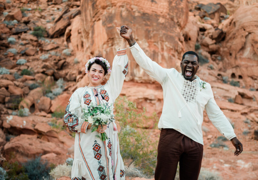 A smiling couple in embroidered clothing stands in a rocky desert landscape, holding hands up in celebration. The woman holds a bouquet and wears a flower crown.