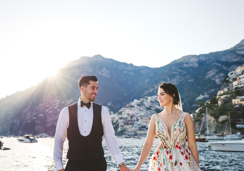 A couple stands hand in hand by the water, dressed in wedding attire. The groom wears a black tuxedo, and the bride wears a floral gown. They look at each other lovingly with a picturesque coastal town and mountains in the background. Boats float nearby.