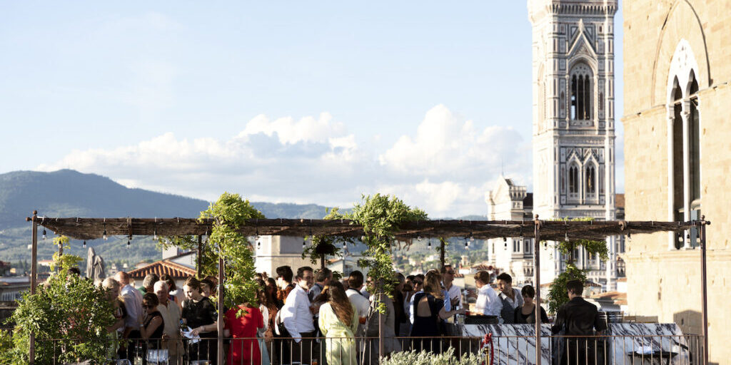A group of people gather on a rooftop terrace with vines overhead, socializing with drinks. A tall stone tower stands in the background on a clear day.