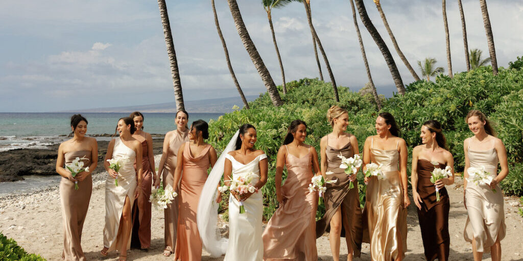 A bride in a white dress walks on a beach with eight bridesmaids in assorted neutral dresses, all holding bouquets, with palm trees and ocean in the background.