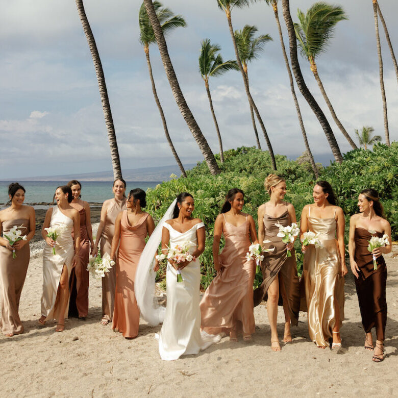 A bride in a white dress walks on a beach with eight bridesmaids in assorted neutral dresses, all holding bouquets, with palm trees and ocean in the background.