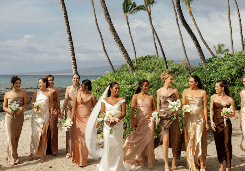 A bride in a white dress walks on a beach with eight bridesmaids in assorted neutral dresses, all holding bouquets, with palm trees and ocean in the background.
