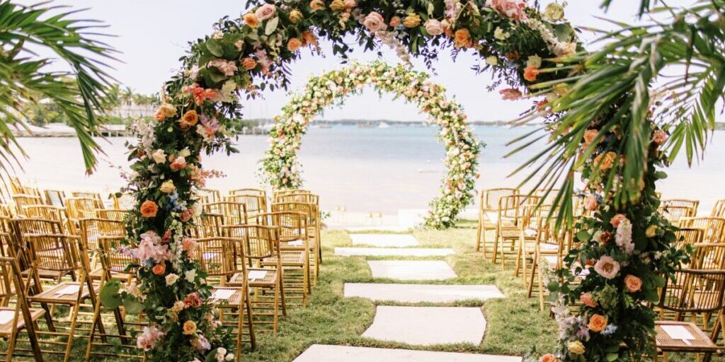 Tropical outdoor wedding setup with floral arches over a grassy aisle, rows of wooden chairs, and a beach backdrop.