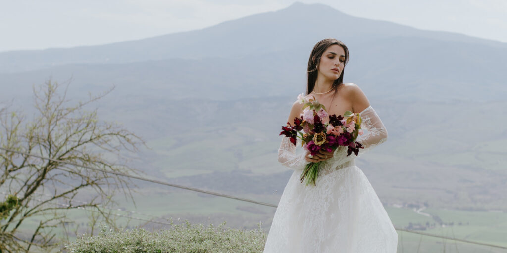A woman in a white wedding dress holds a bouquet of flowers, standing outdoors with mountains and greenery in the background.